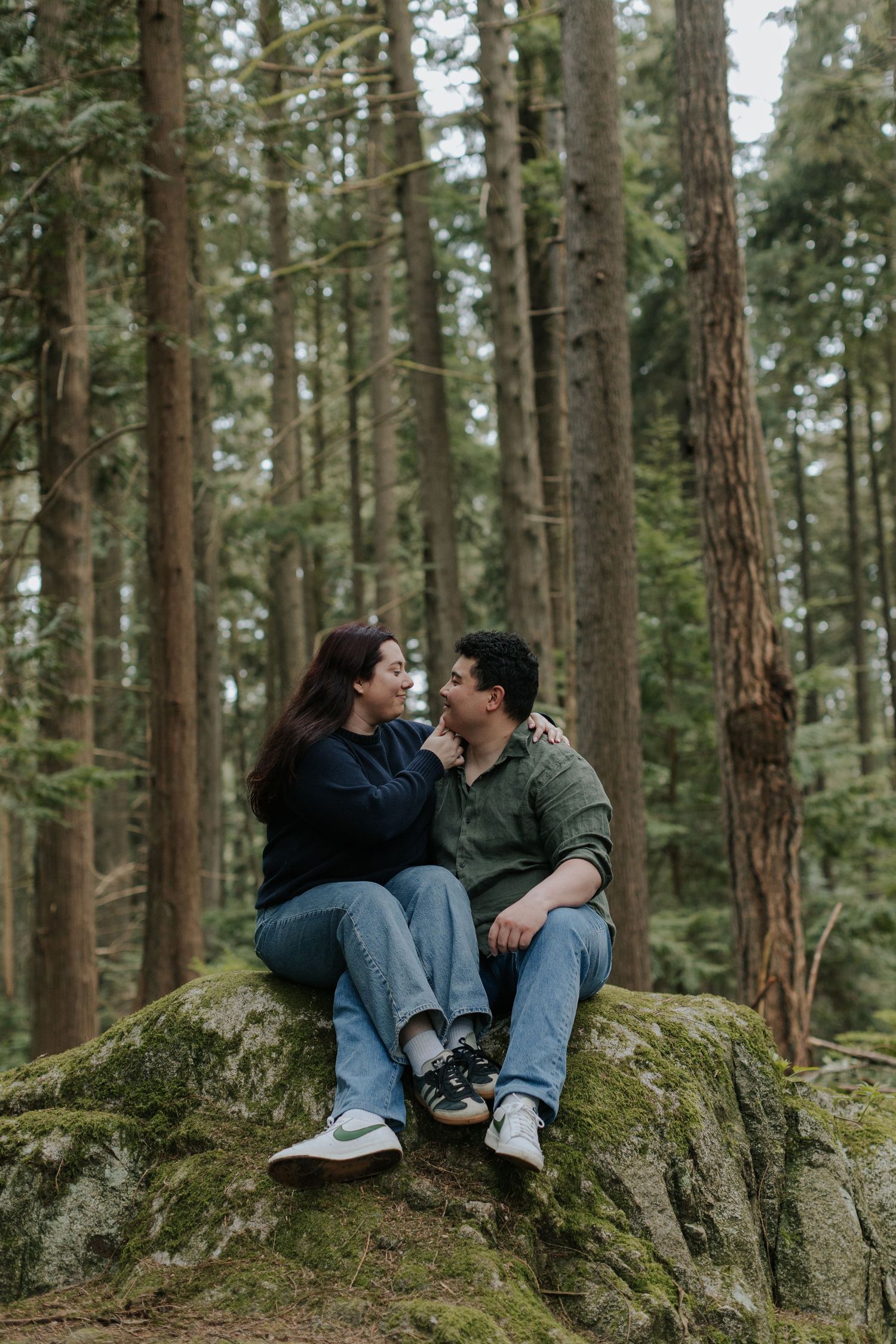 Couple sitting on a mossy rock in a forest, gazing at each other romantically among tall evergreen trees.