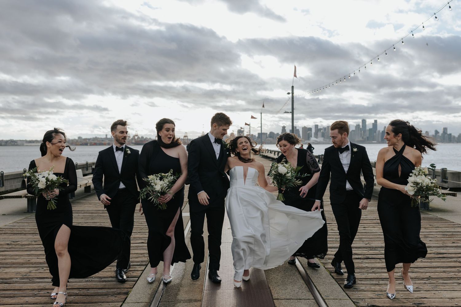 Wedding party members in black and white attire running playfully along a waterfront boardwalk.