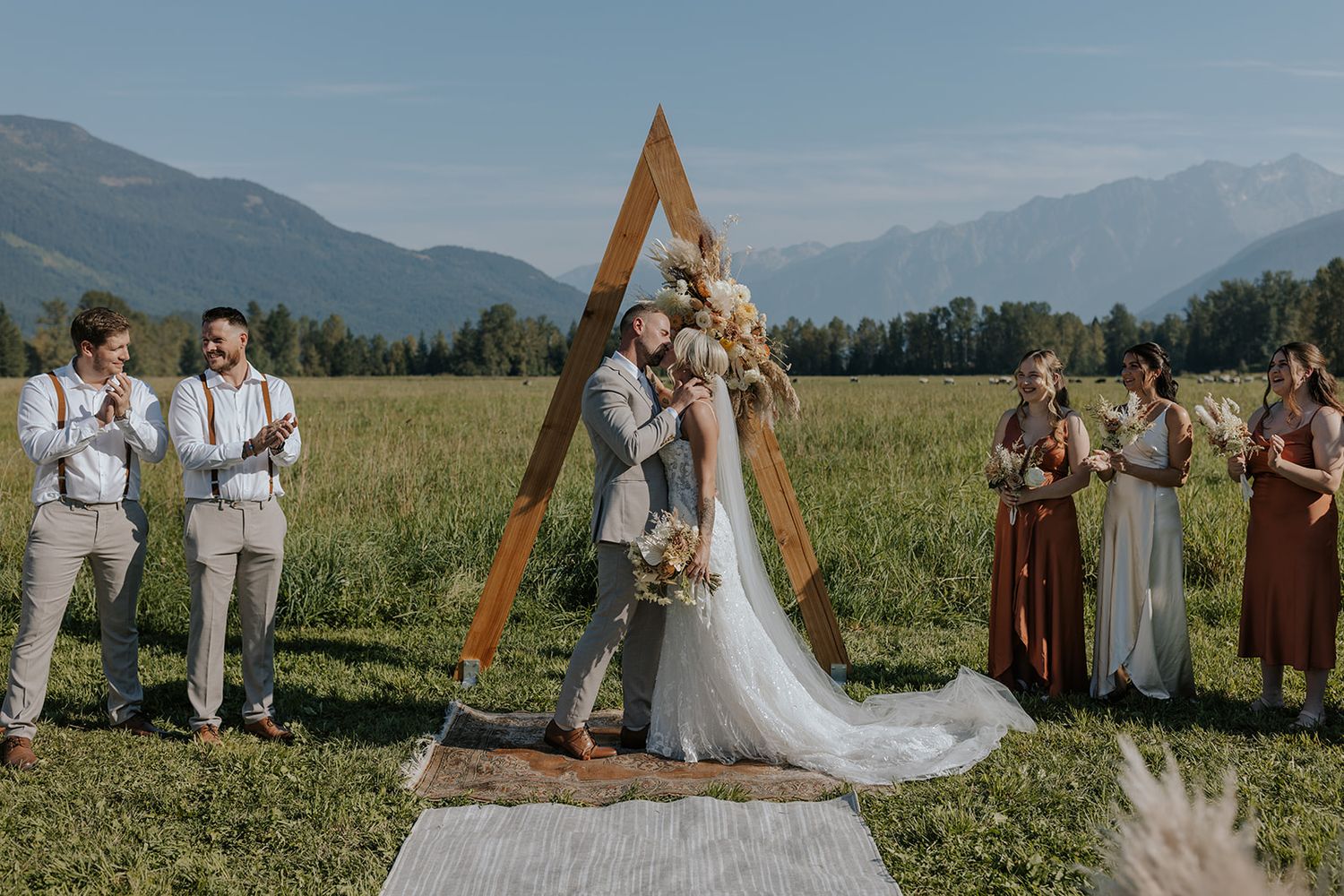 A wedding ceremony takes place under a wooden arch in a scenic mountain meadow with wildflowers and distant peaks.