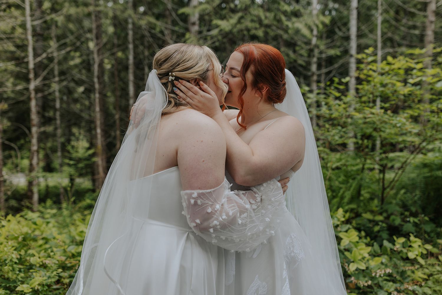 Two brides in white dresses embrace tenderly in a wooded outdoor setting.