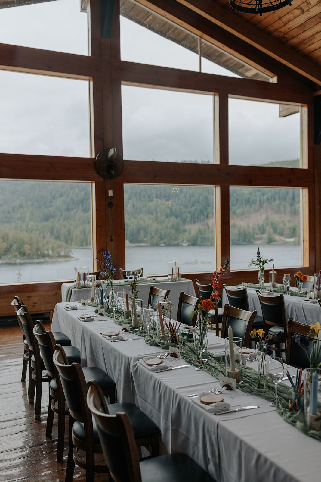 Wedding reception tables set with white linens and greenery against large windows overlooking a lake.