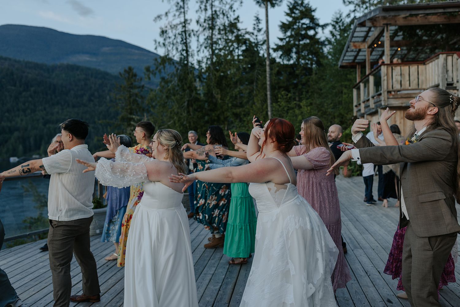 Outdoor wedding celebration with guests dancing on a deck surrounded by mountain scenery.