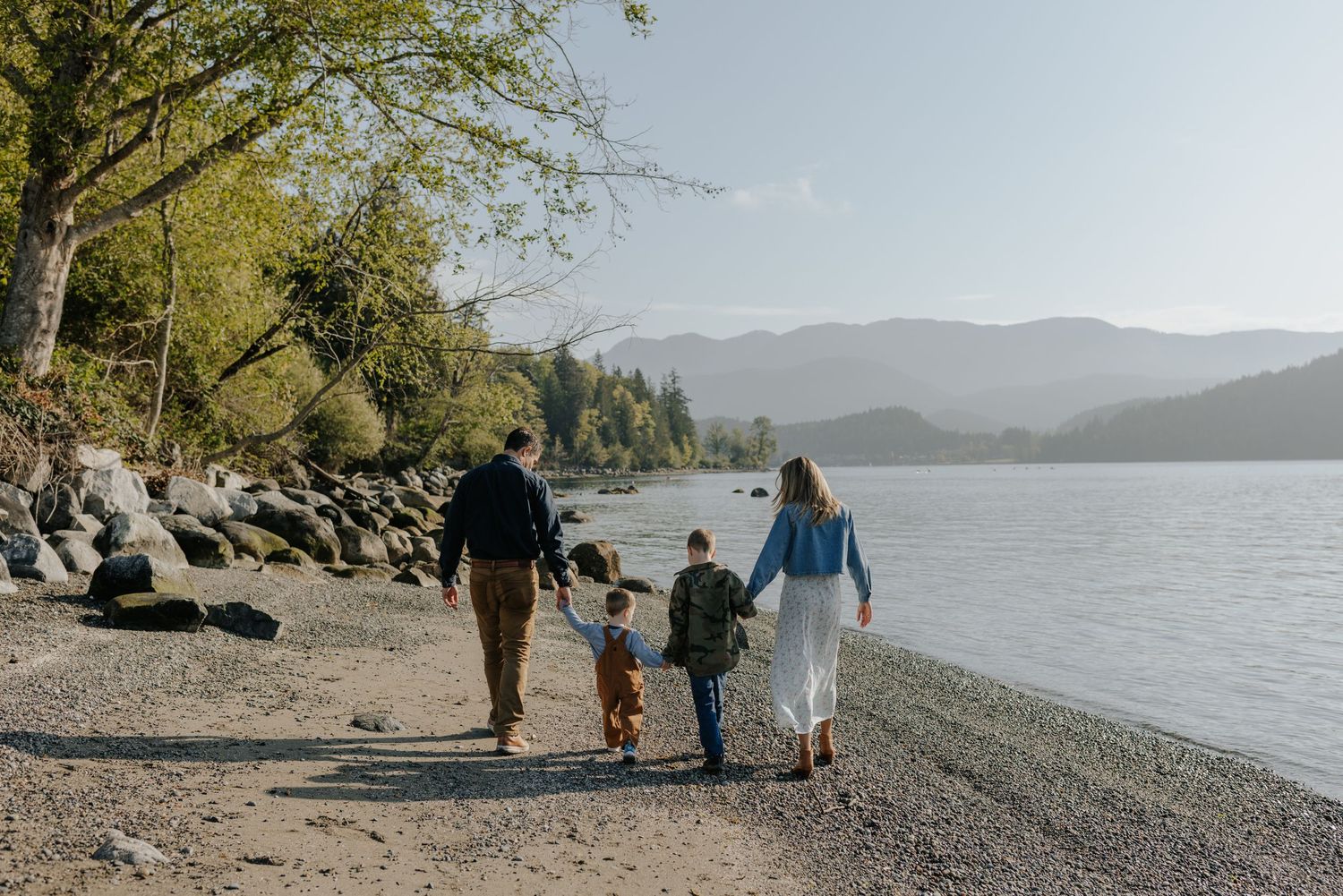 Family of four walking hand-in-hand along a rocky lakeside shore with mountains in the background.