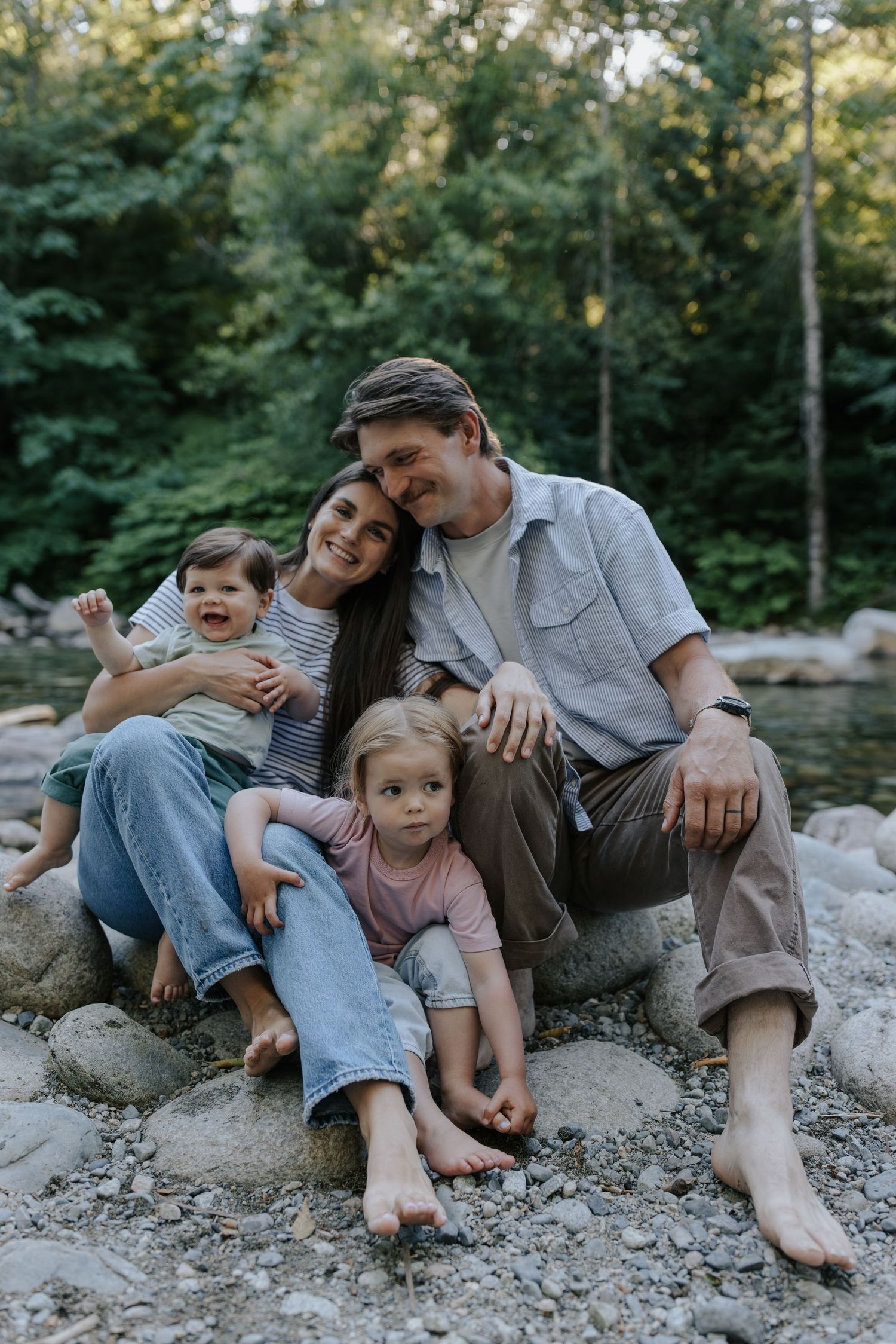 A happy family of four sits barefoot on rocks by a forest stream, parents cuddling two young children outdoors.