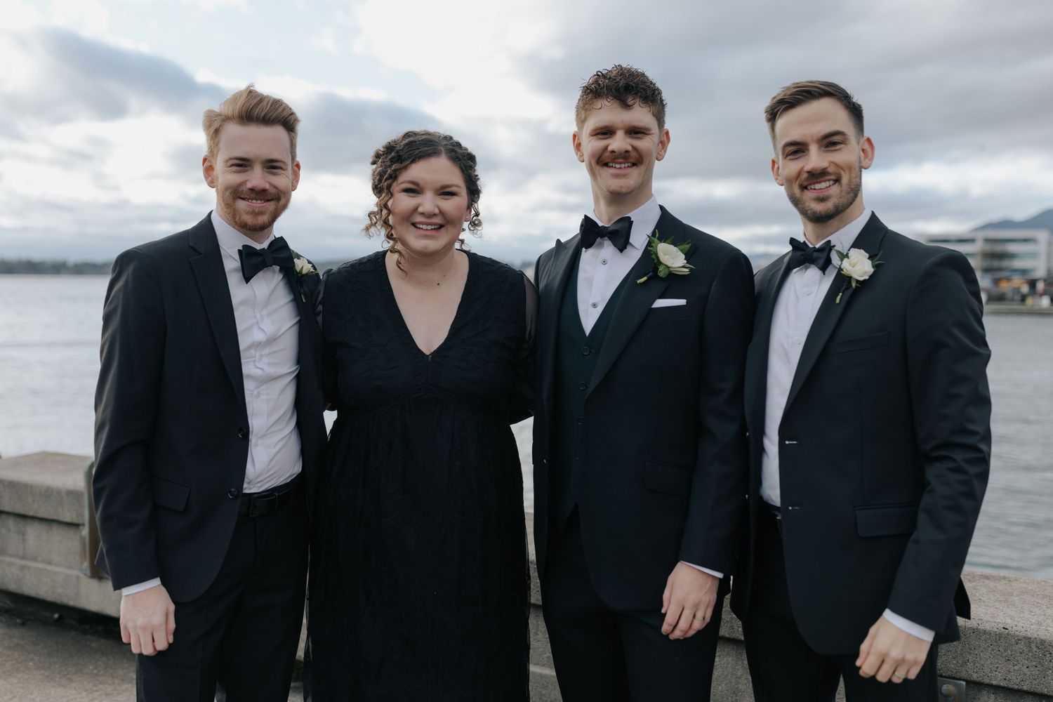 Groomsmen in black tuxedos smile together during outdoor wedding photos by the water.