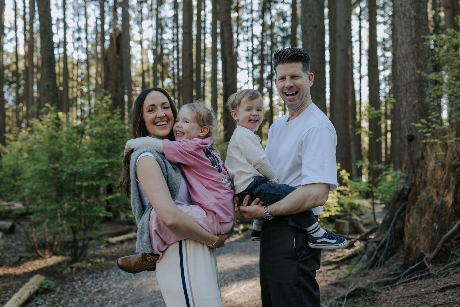 Happy family of four posing in a forest, mother carrying baby in pink wrap, father holding toddler, smiling outdoors.