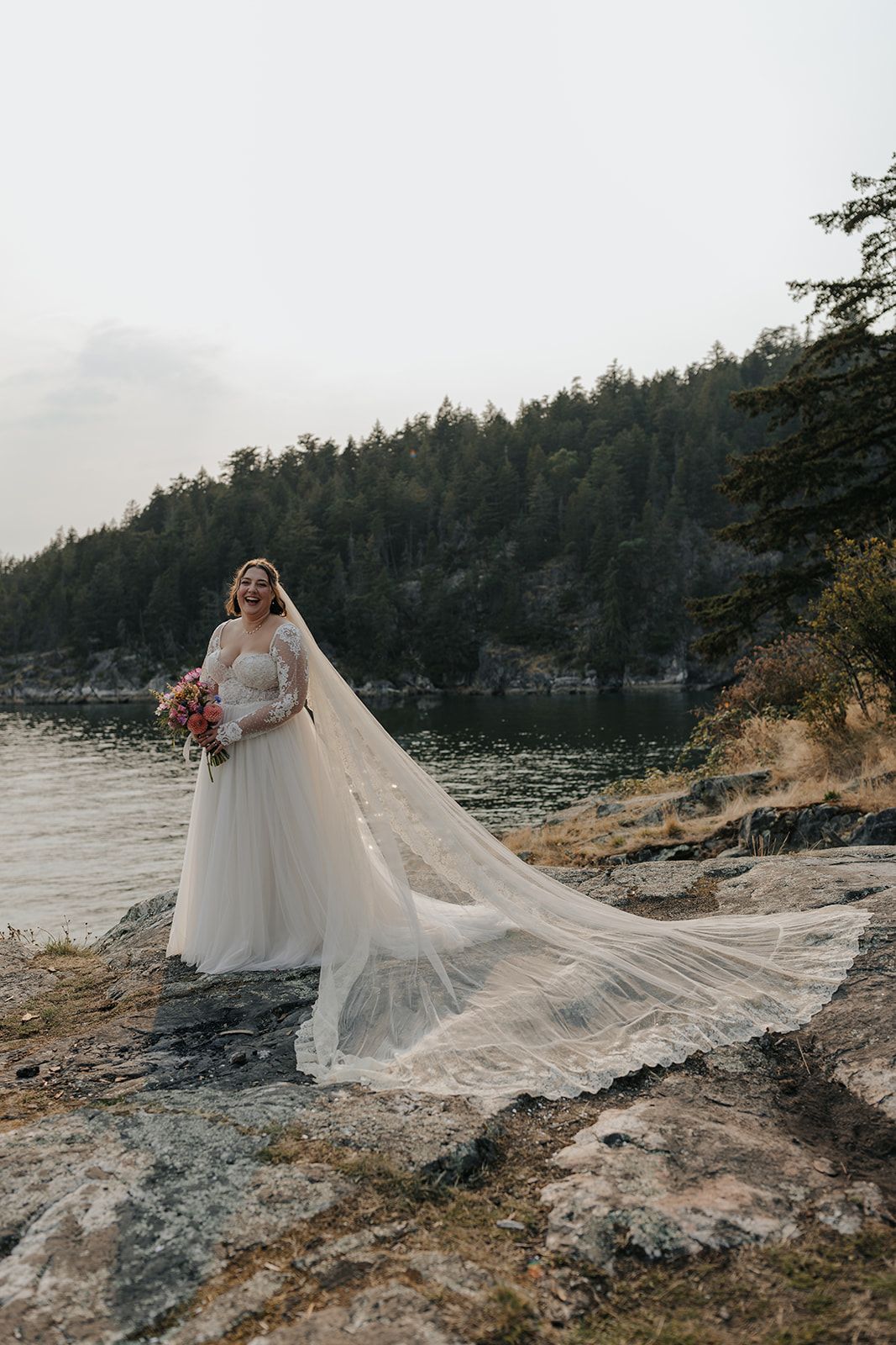 A bride in a flowing dress and veil poses against a dramatic coastal landscape.