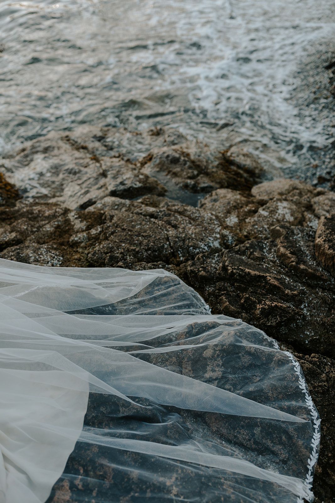 White wedding veil flowing over rocky coastal terrain near water.