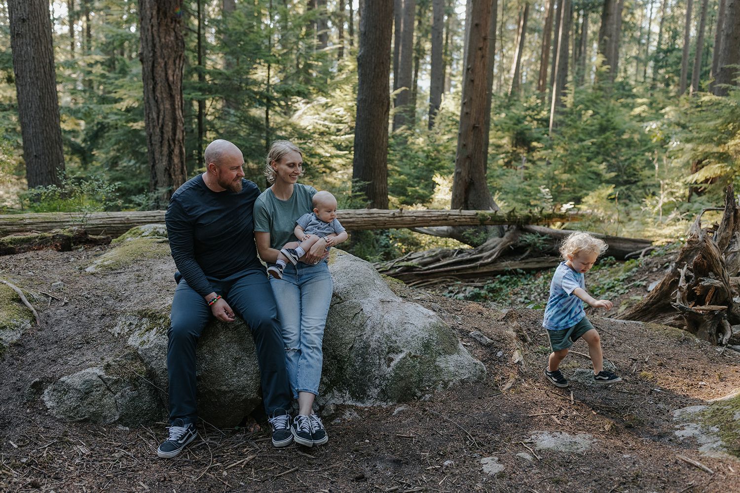 Family relaxing on large boulder in forest while child explores nearby.