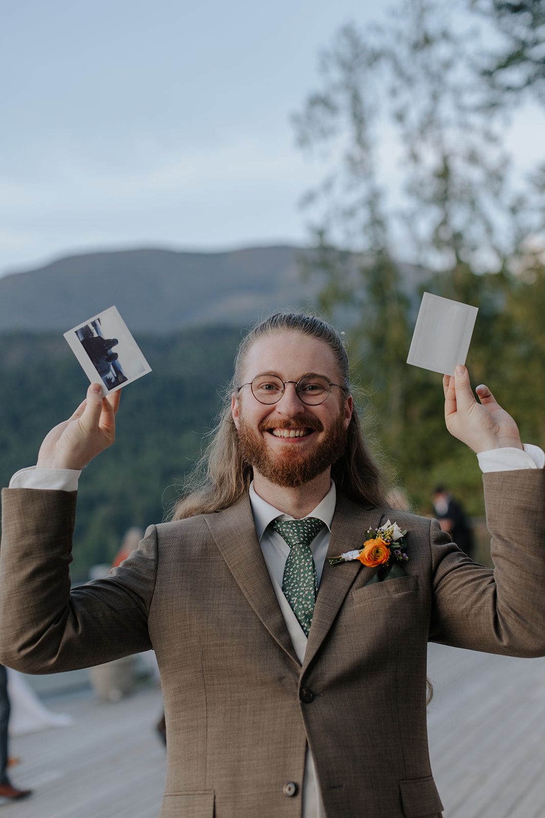 Person in brown suit holding up polaroid photos with joyful expression on outdoor deck.