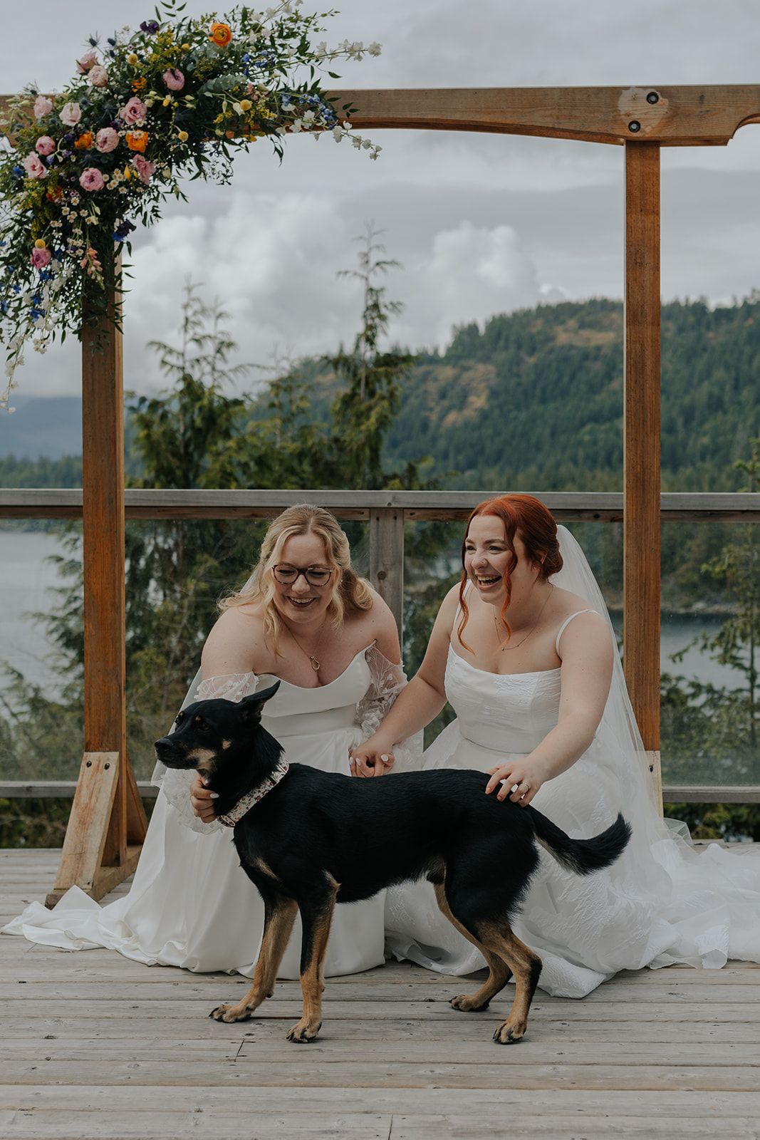 Two brides in white dresses play with their dogs during wedding photos on a scenic deck.