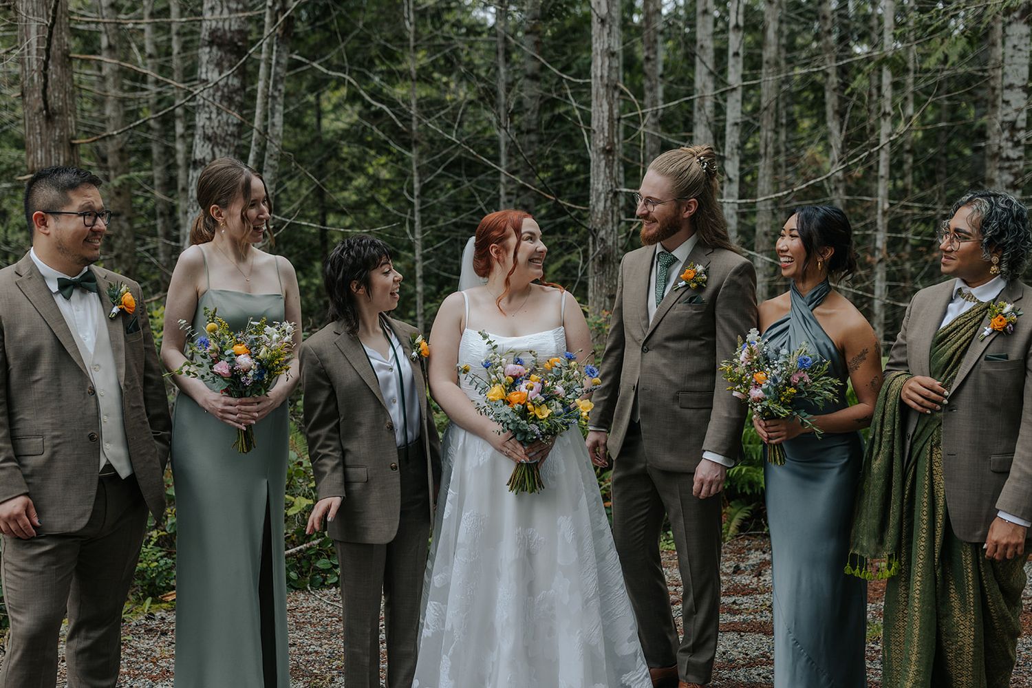 Wedding party poses together in a forest setting with bridesmaids in sage green dresses and colorful bouquets.