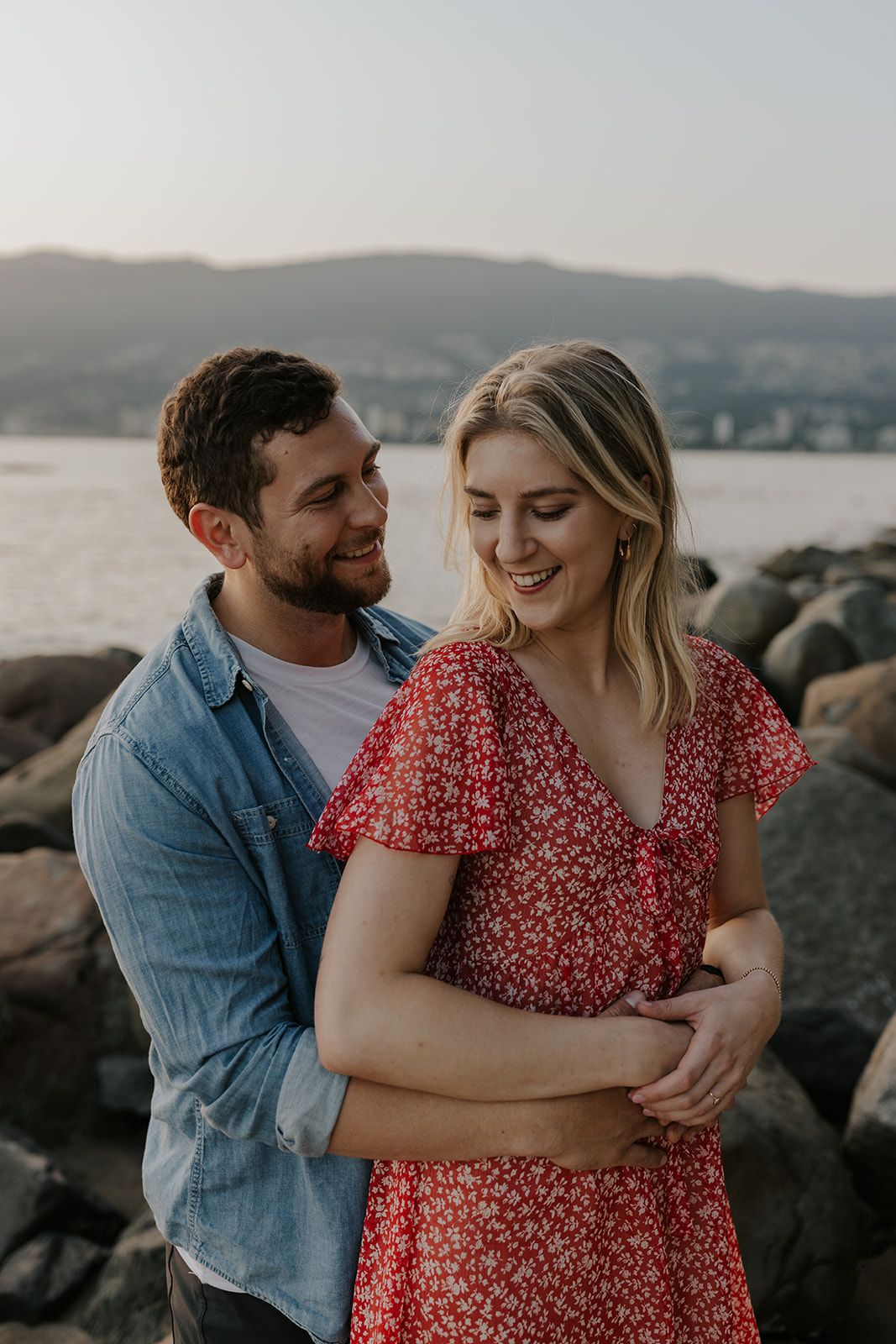 A couple embracing on rocky coastline during sunset with mountains in background.