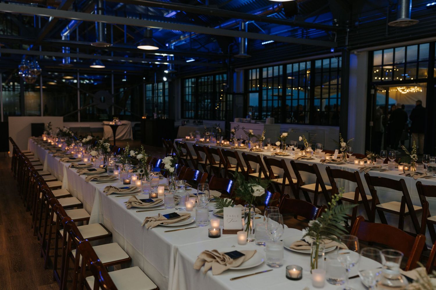 Long banquet tables set for an elegant wedding reception with white linens and greenery in an industrial venue.