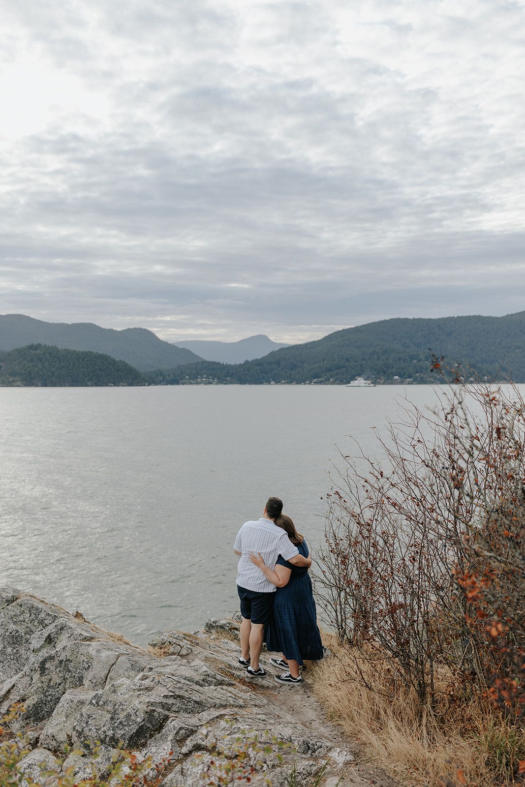 A couple sits together on coastal rocks overlooking a misty mountain bay.