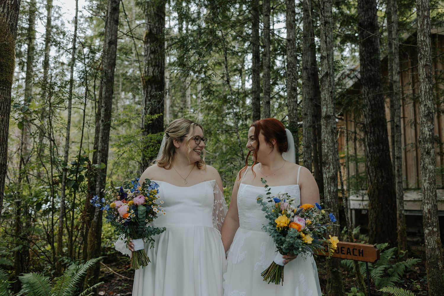 A wedding couple in matching white gowns poses together on a forest path holding vibrant floral bouquets.