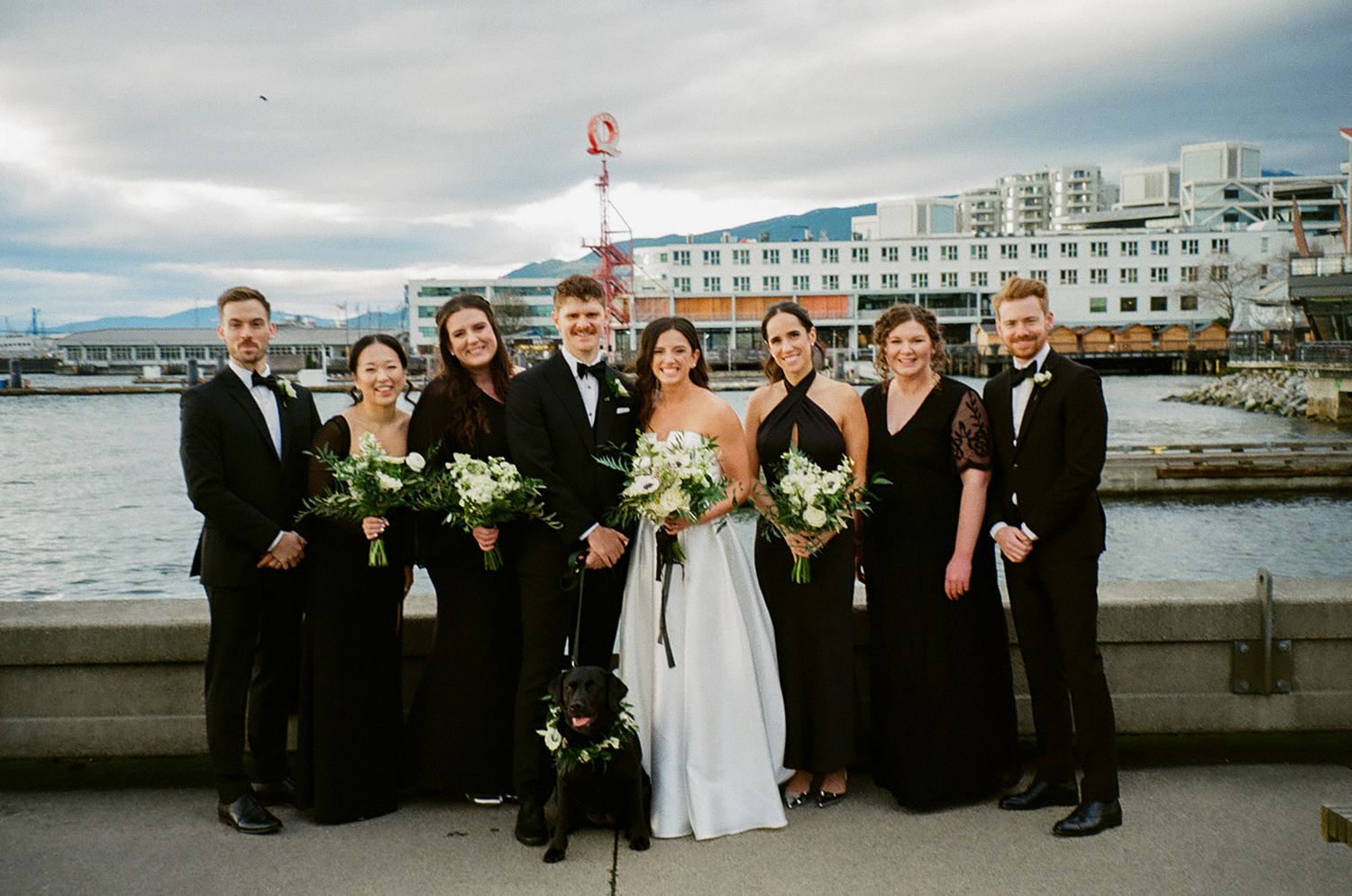 Wedding party poses together on waterfront with city buildings in background.