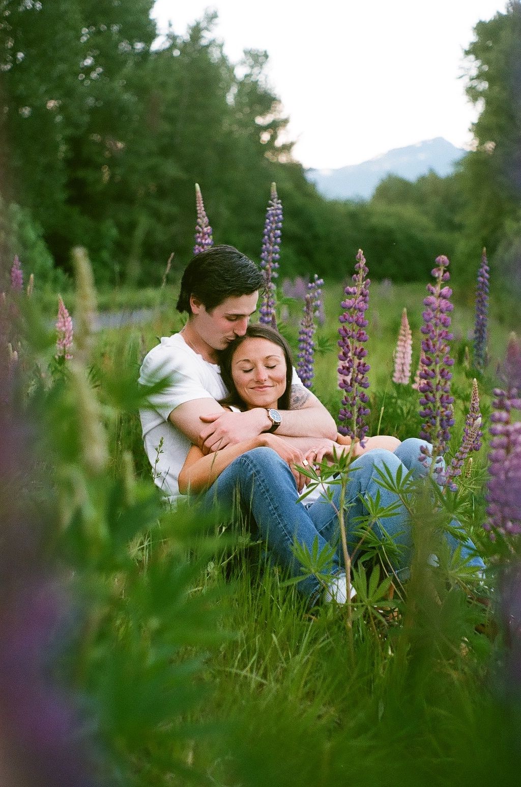 A romantic couple embraces in a field of purple lupines with mountains in the background.