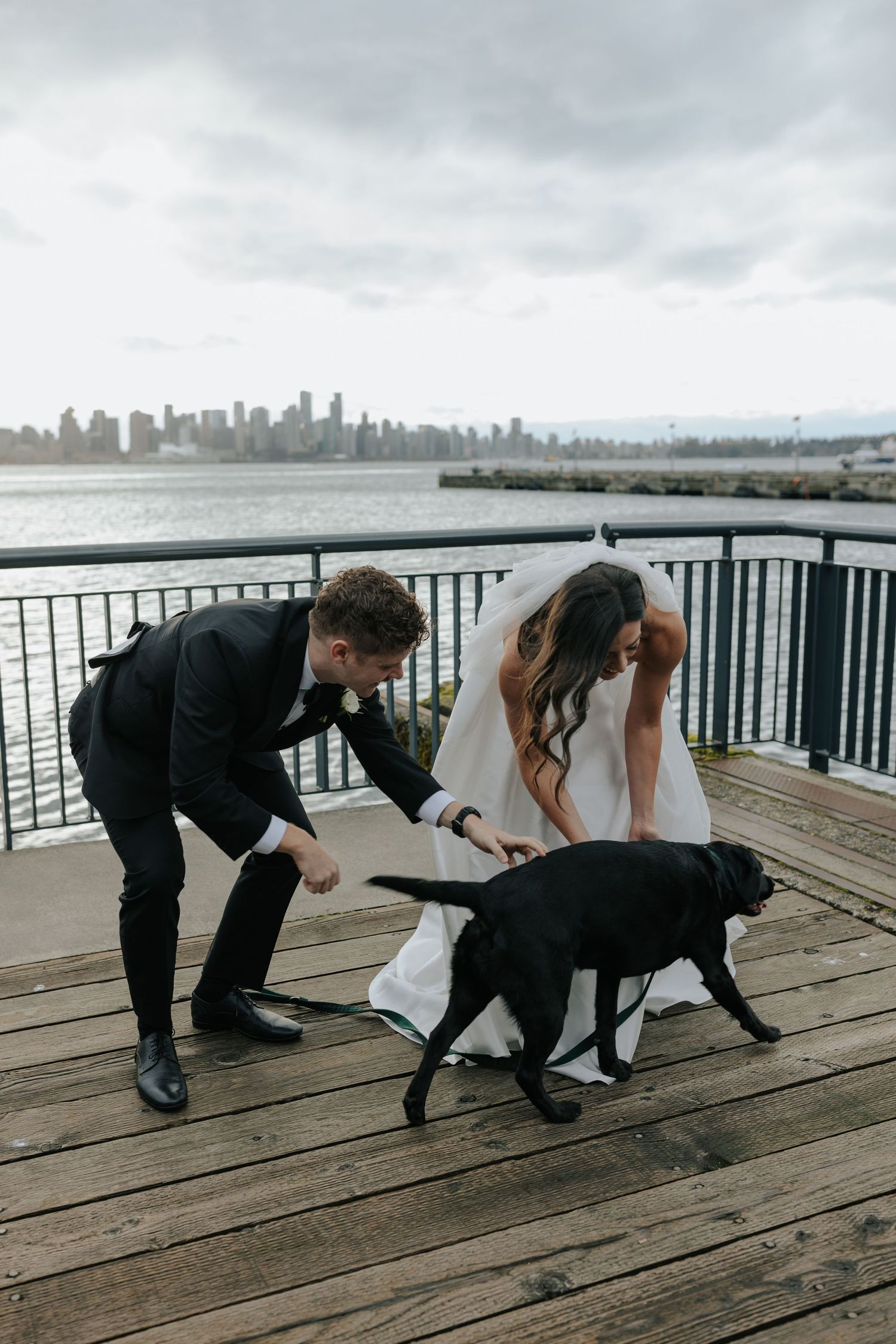 A couple and their black dog walk along a wooden pier with the city skyline visible in the background.