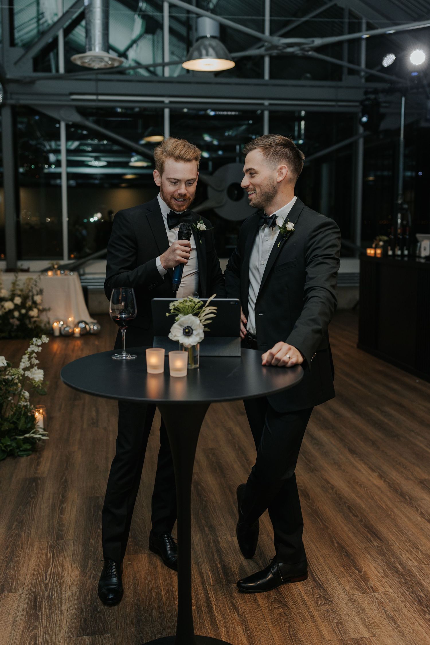 Groomsmen in black tuxedos sharing drinks and conversation on wooden dance floor.