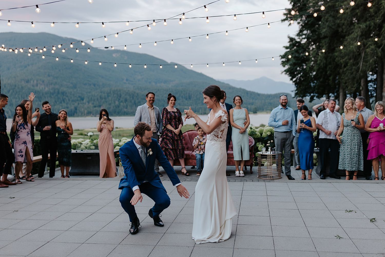 Outdoor wedding reception with string lights and dancing guests on a patio overlooking mountain views at sunset.