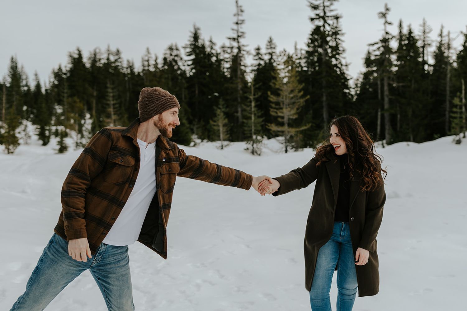 A couple plays in the snow surrounded by evergreen trees on a winter mountainside.