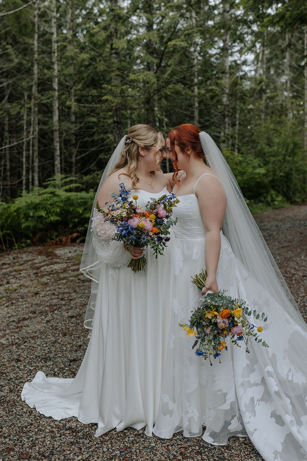 Two brides with colorful bouquets share an intimate moment surrounded by towering forest trees.
