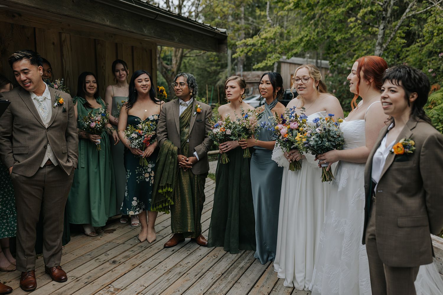 Wedding party in green dresses and suits gathered on wooden deck for group photos.