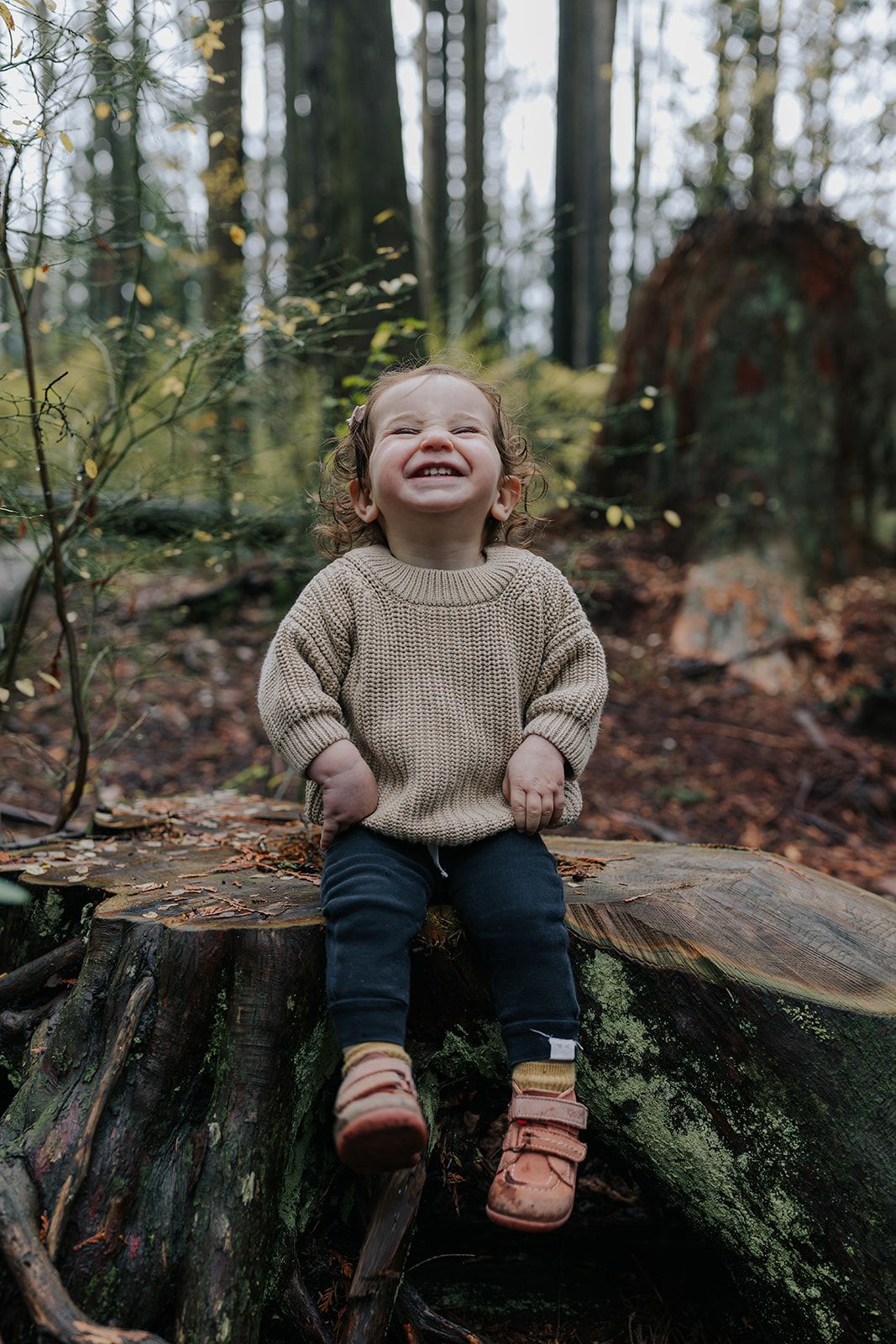 Child in knit sweater sitting and laughing on mossy tree stump in forest.