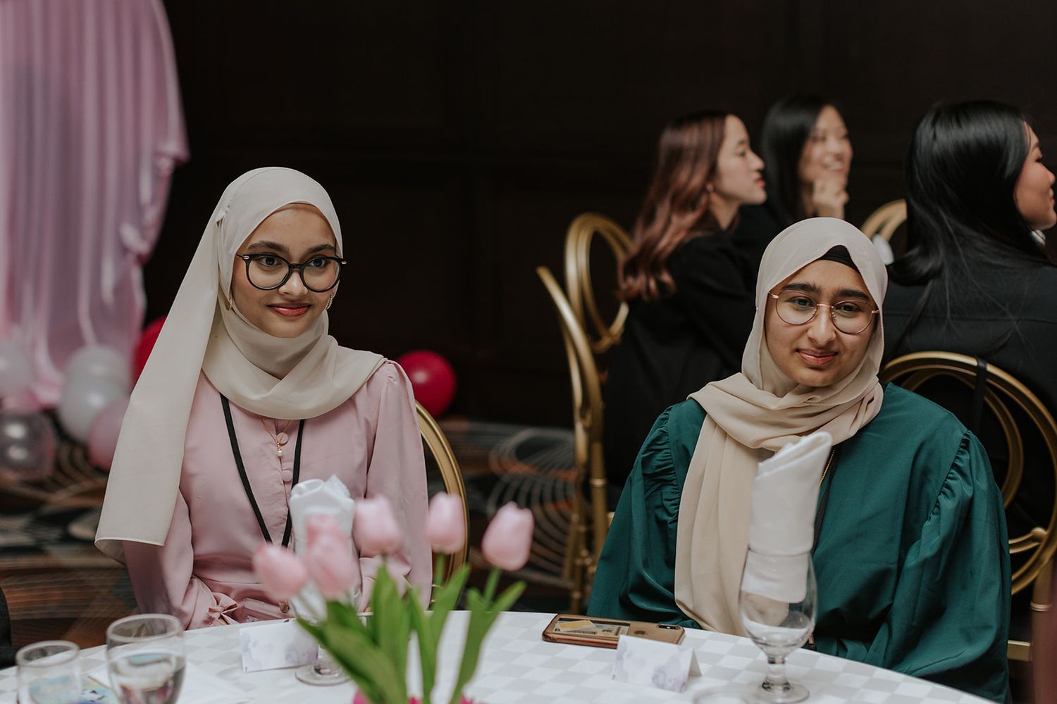 Two women in hijabs wearing pink and green outfits smile while seated at a table during an event.