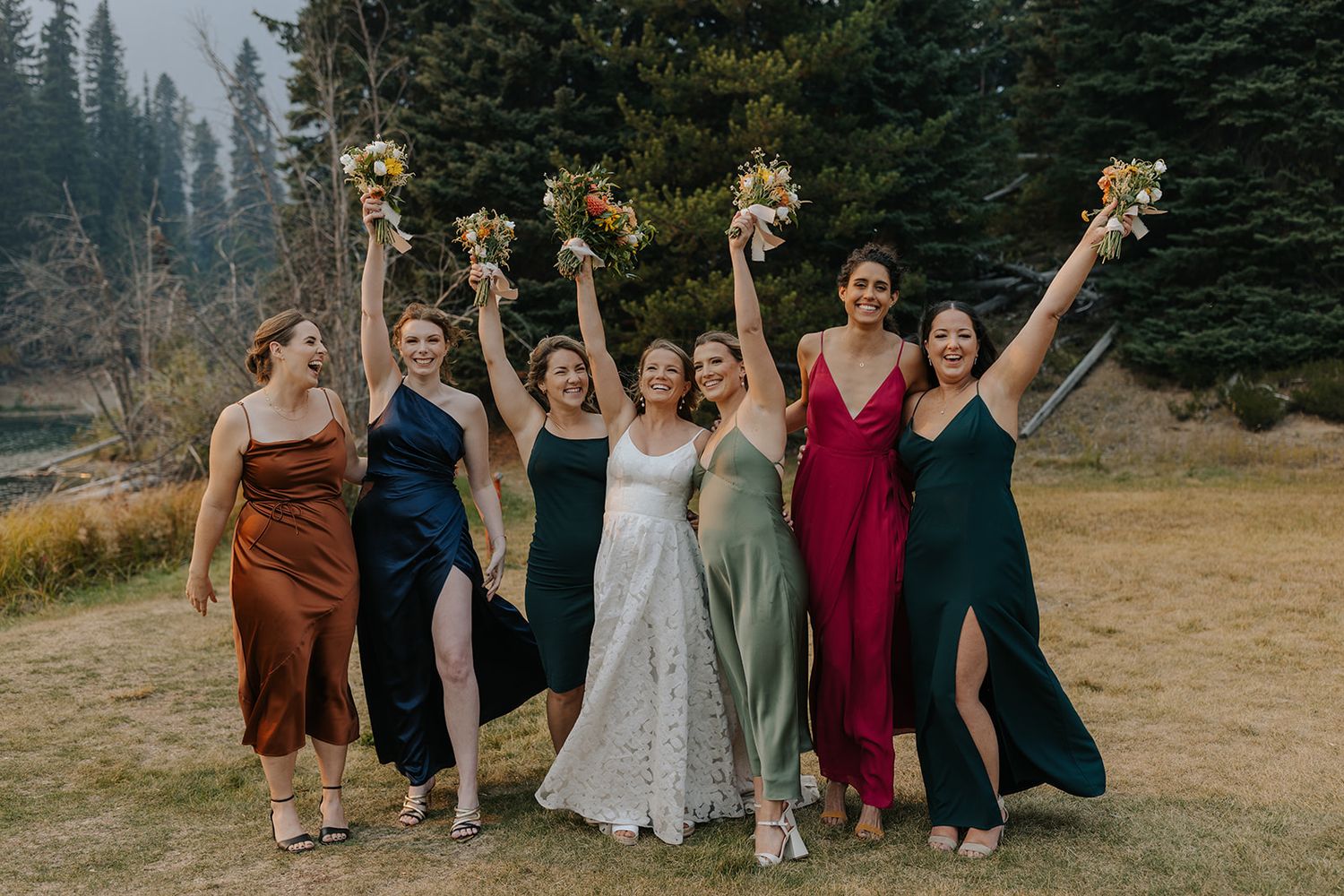 Wedding party in mix of burgundy and earth tone dresses posing outdoors with evergreen trees.