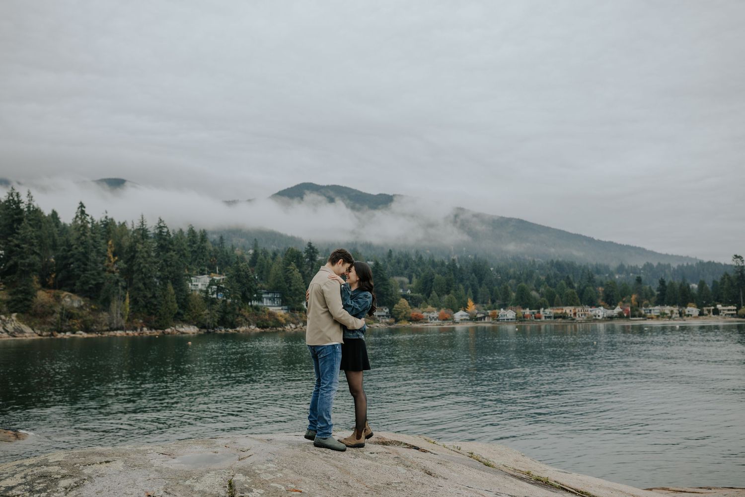 Couple embracing on rocky shoreline with misty mountains and evergreen trees reflected in calm water on overcast day.