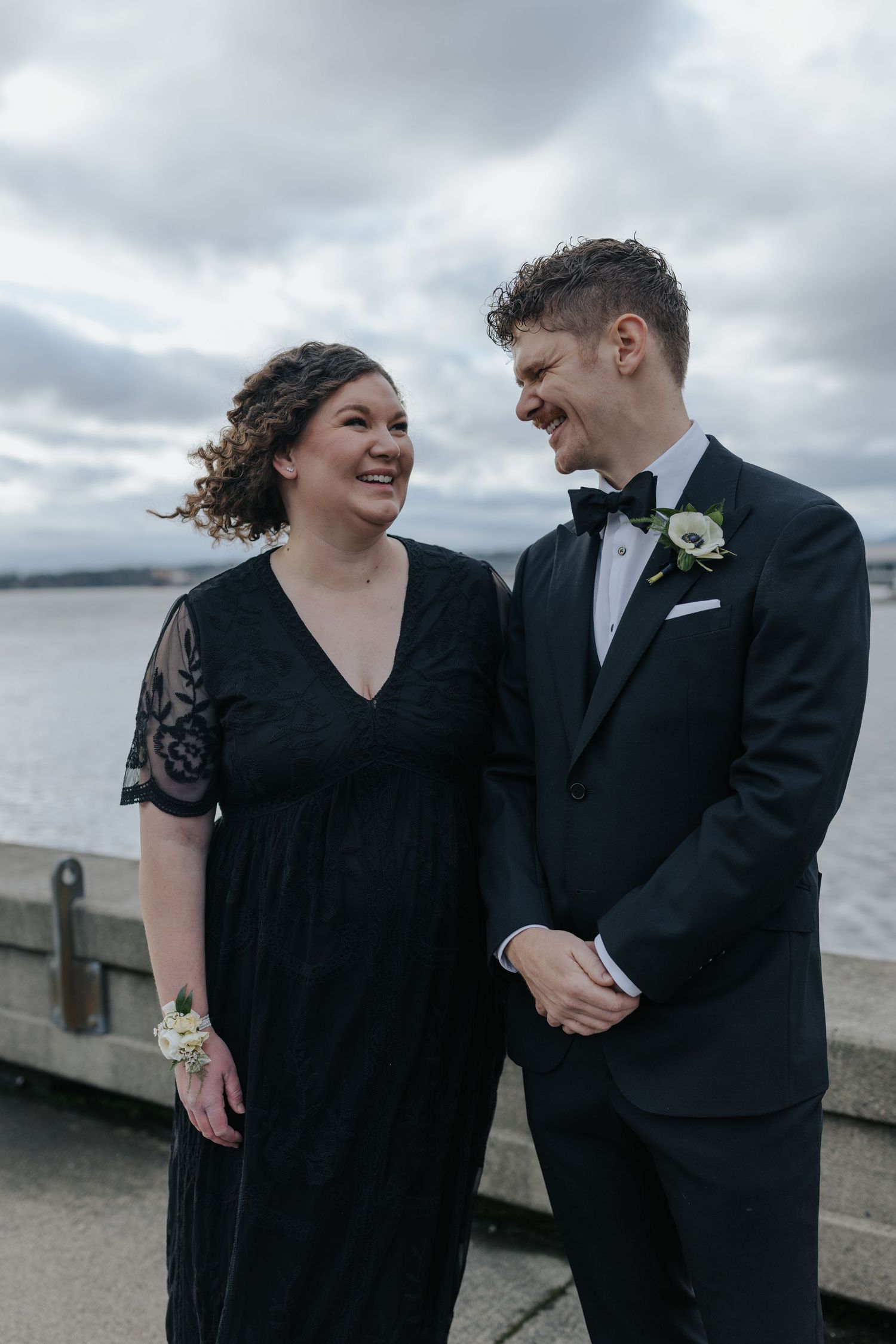 A couple shares a joyful moment together by the waterfront on their wedding day under moody cloudy skies.