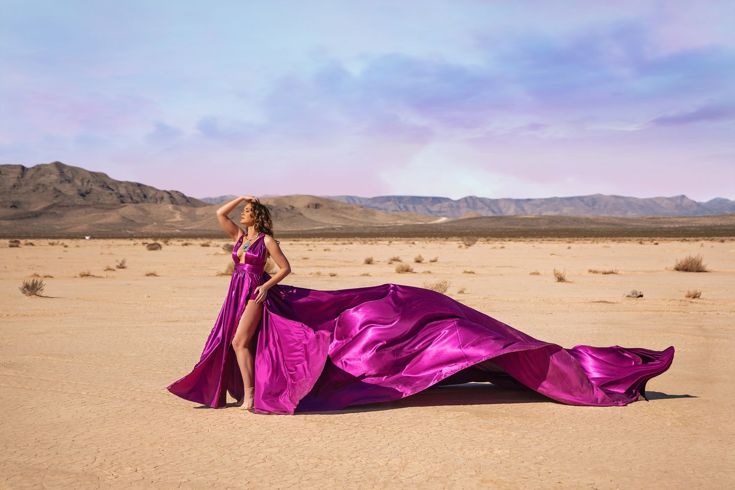 A vibrant magenta gown billows dramatically across a desert landscape under a pastel sunset sky.