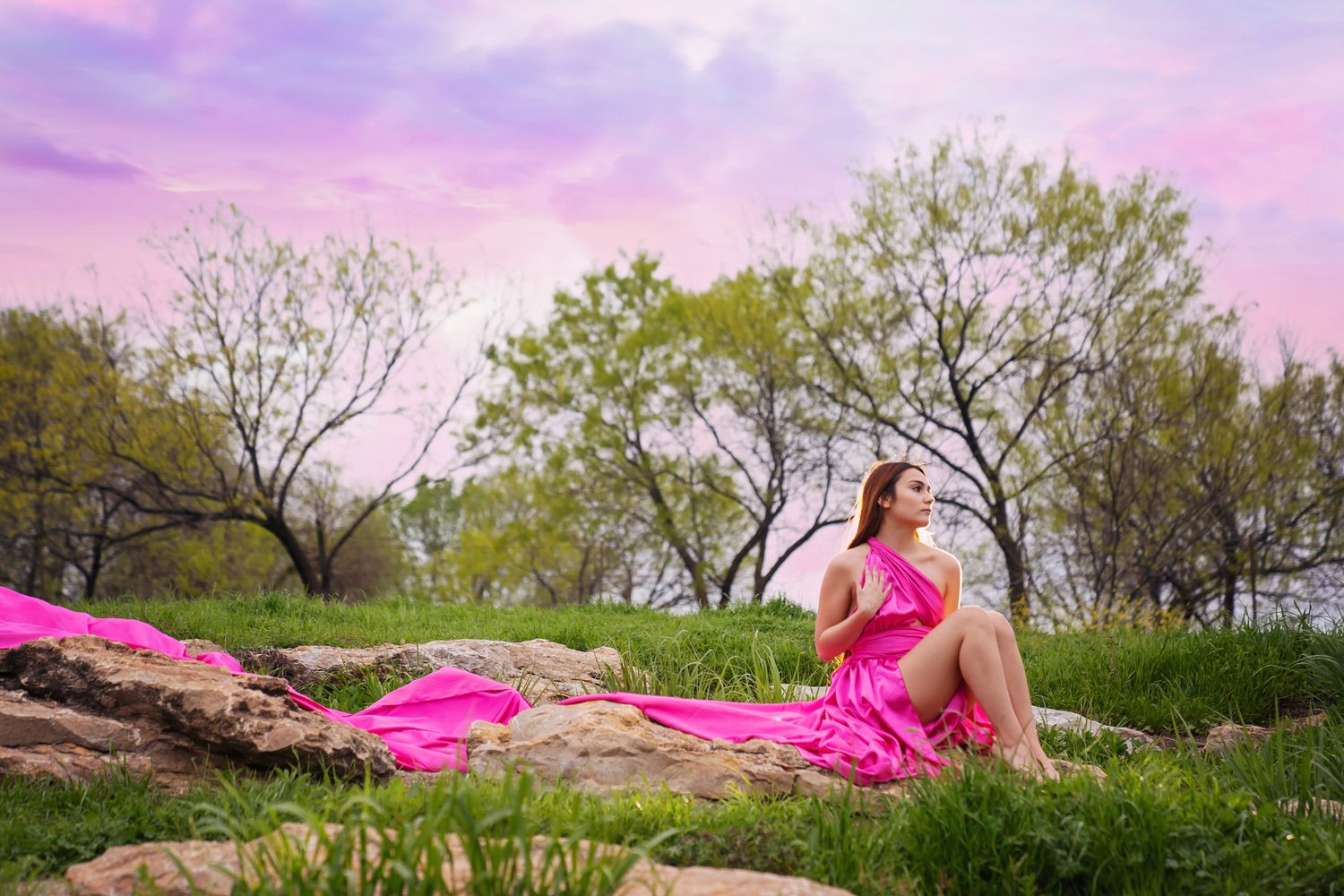 A bright pink gown flows dramatically across large rocks in a natural outdoor setting with pink sunset sky.