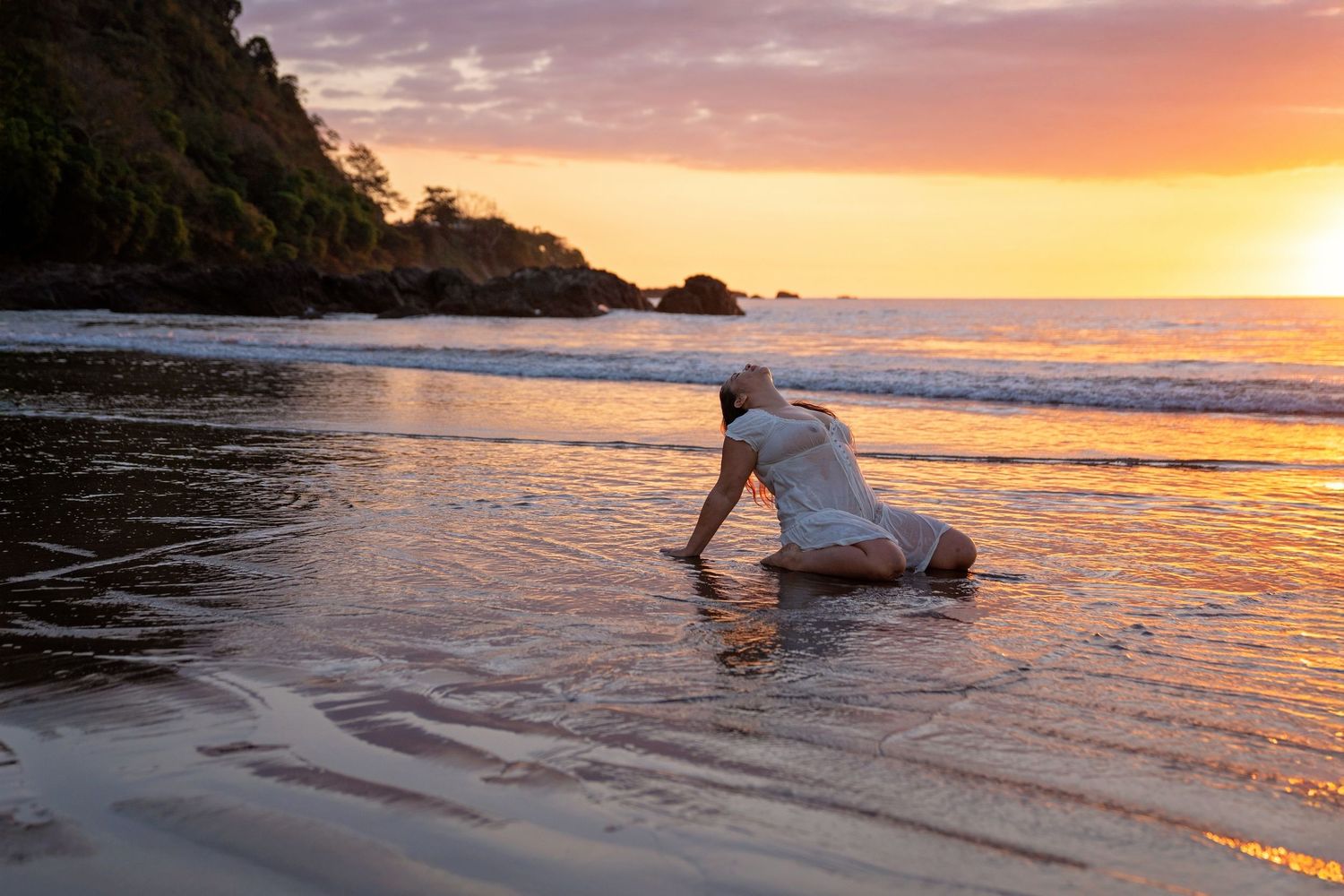 Woman kneeling on a sandy beach touching the water during a vibrant golden sunset with waves and rocky cliffs in background.