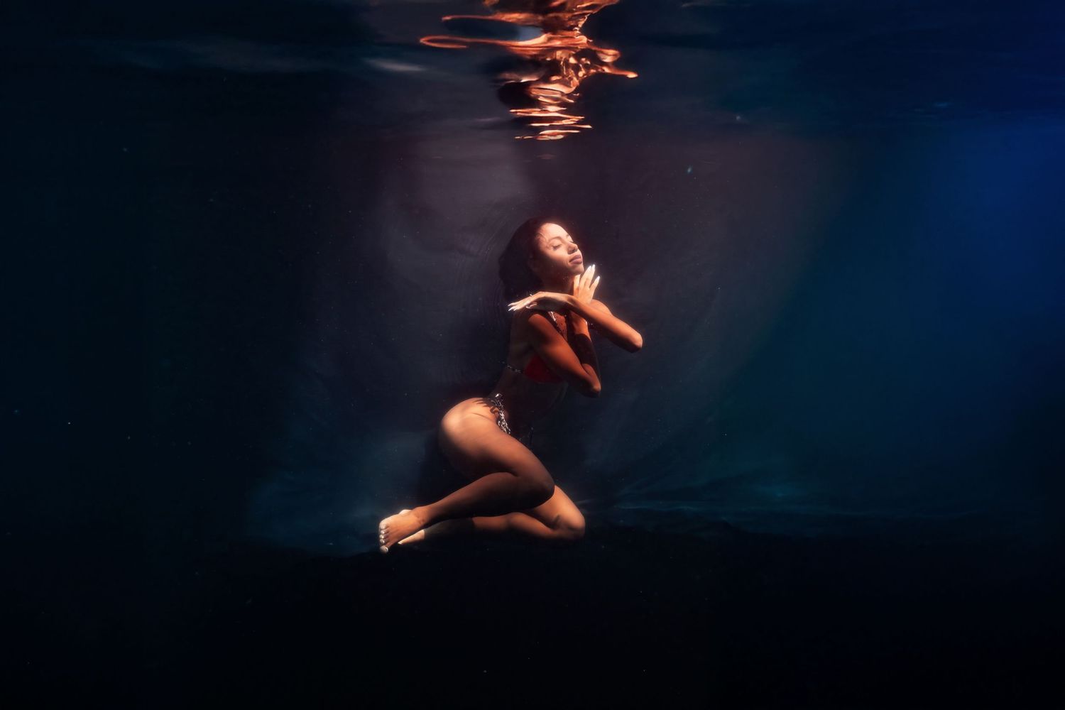 Woman in red swimsuit poses gracefully underwater in dark water, illuminated by light reflecting off the surface.