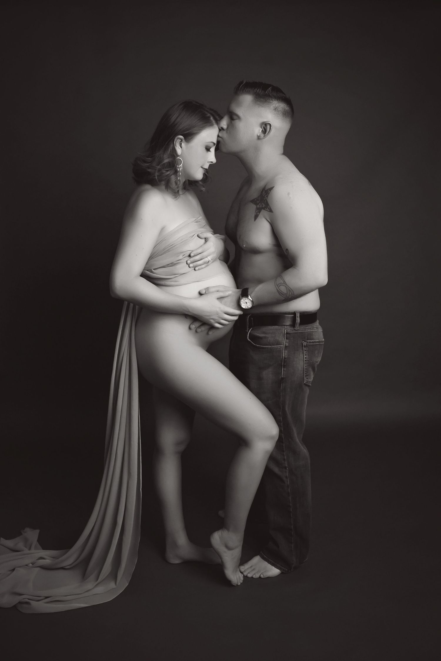 Dramatic studio maternity photo with flowing fabric and intimate couple pose against dark backdrop.
