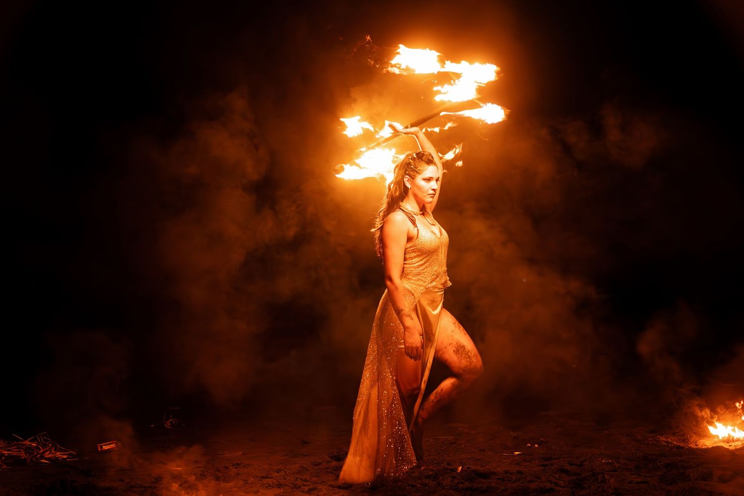 Woman in orange dress performing fire dancing at night with flames and smoke surrounding her.