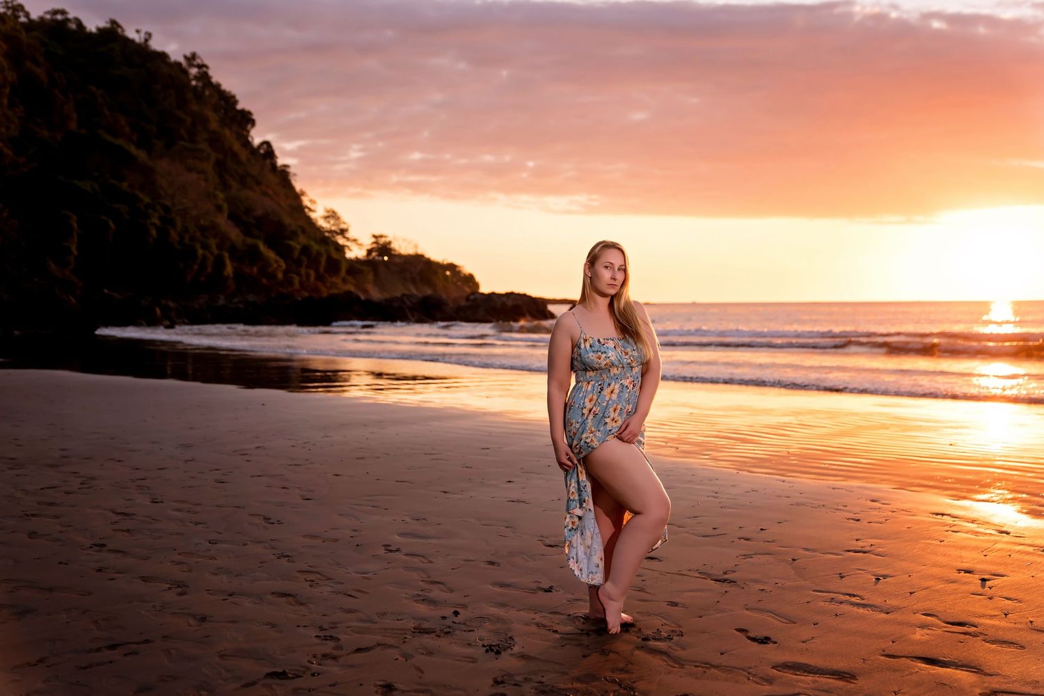 Woman in floral dress posing on sandy beach at golden sunset with ocean waves and lush green cliffs.