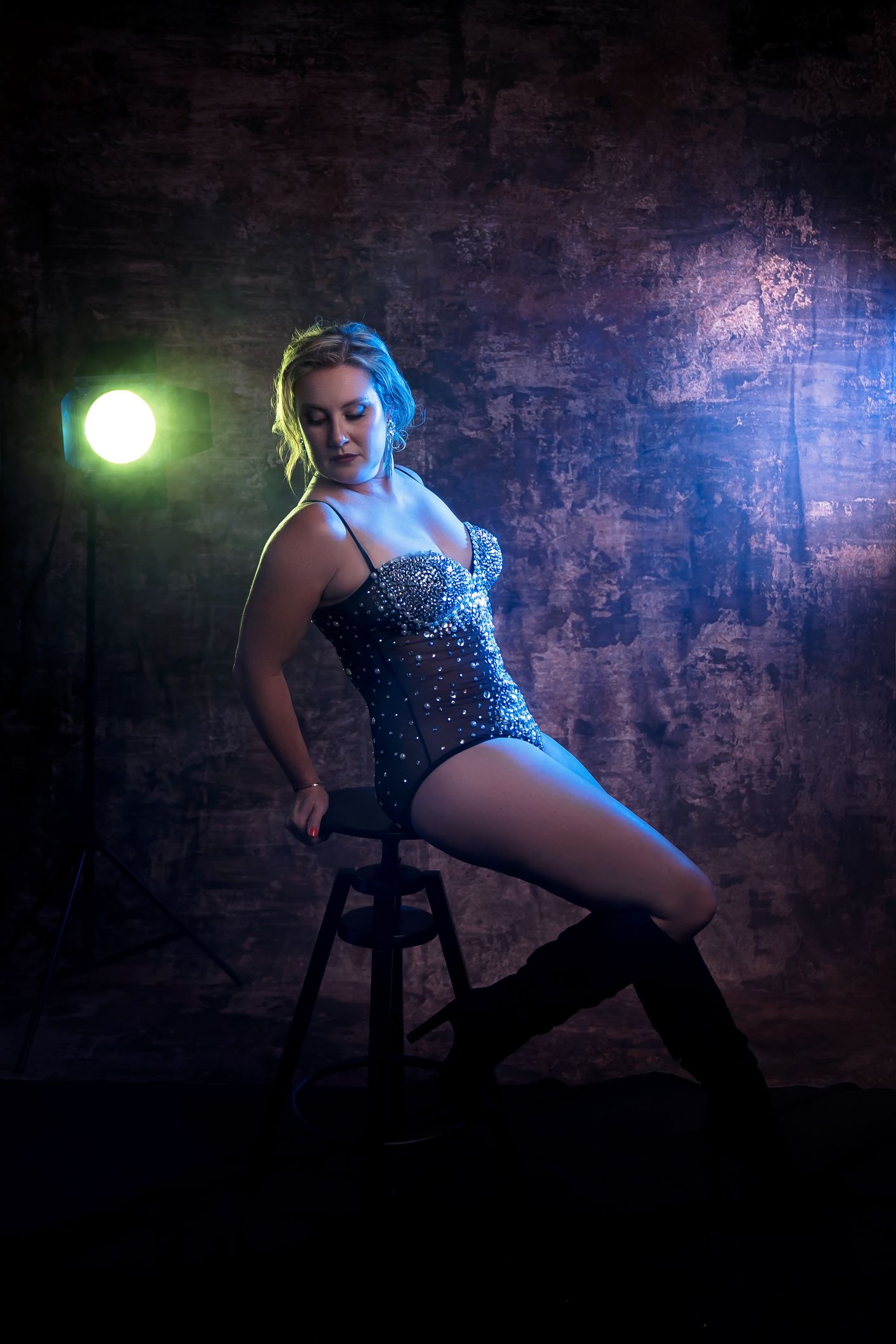 Dramatic boudoir session with sparkly blue bodysuit against brick wall and colored lighting.