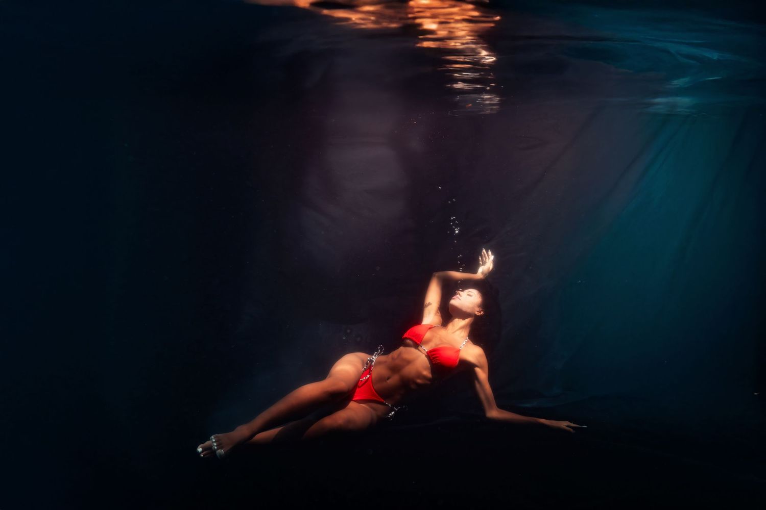 Woman in red bikini swimming underwater with dramatic light rays in dark blue water.