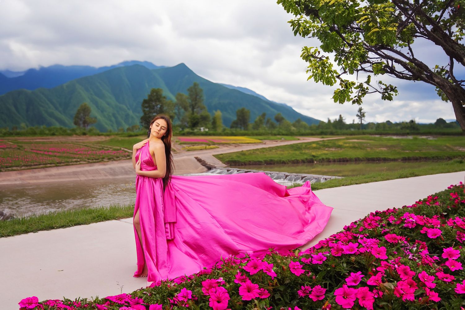 A bright pink dress flows dramatically against a backdrop of mountains and blooming flowers on a scenic pathway.