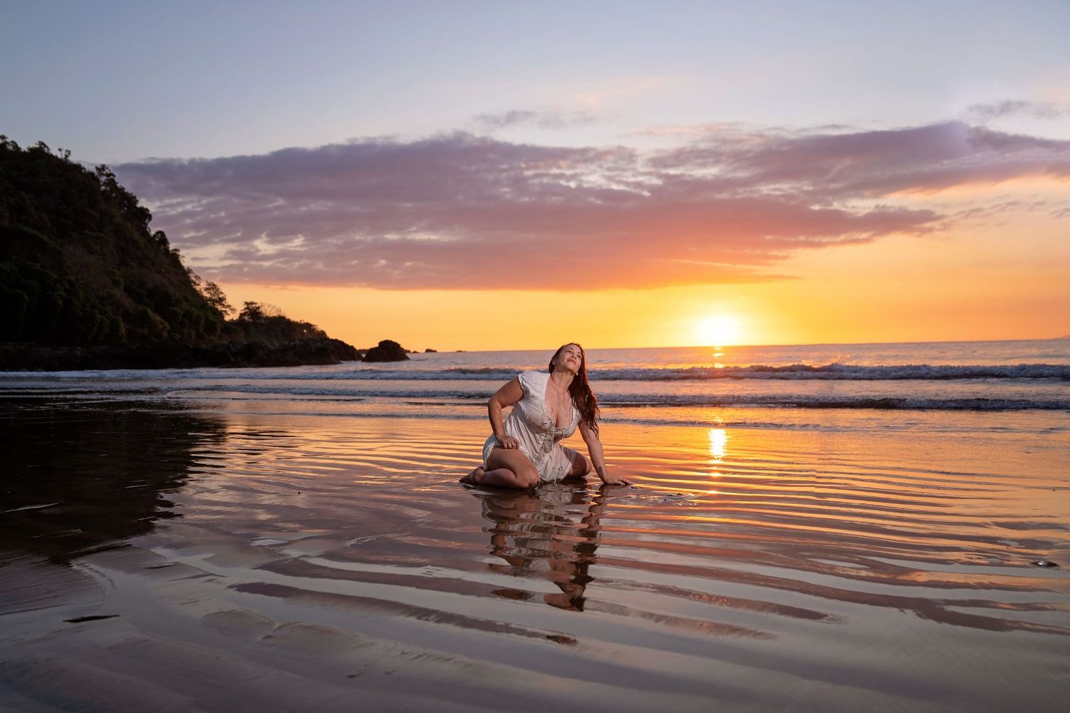 Woman in white dress sitting on reflective beach sand during golden sunset with ocean waves and dramatic sky.