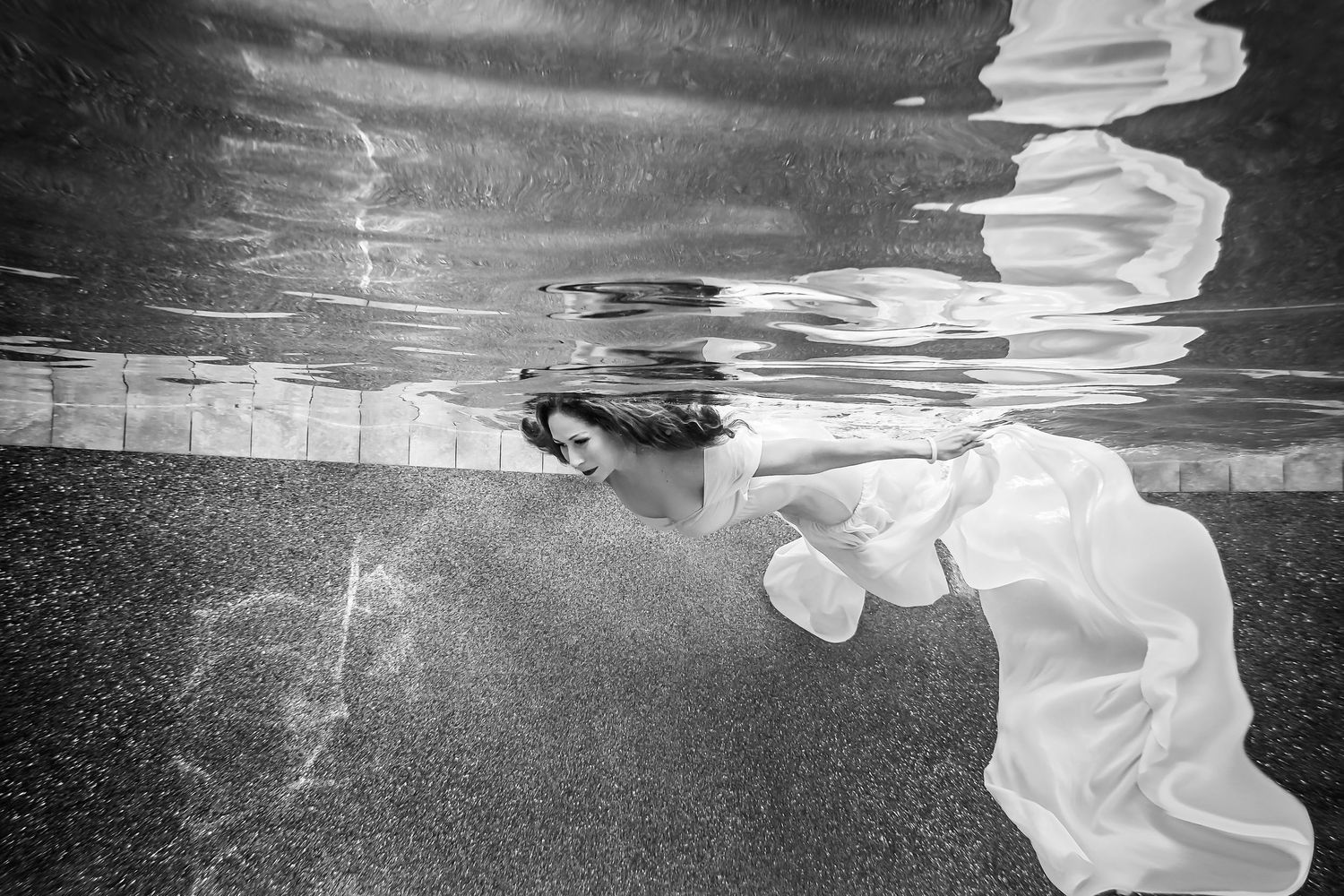 Black and white underwater portrait showing flowing white fabric and rippling water surface.