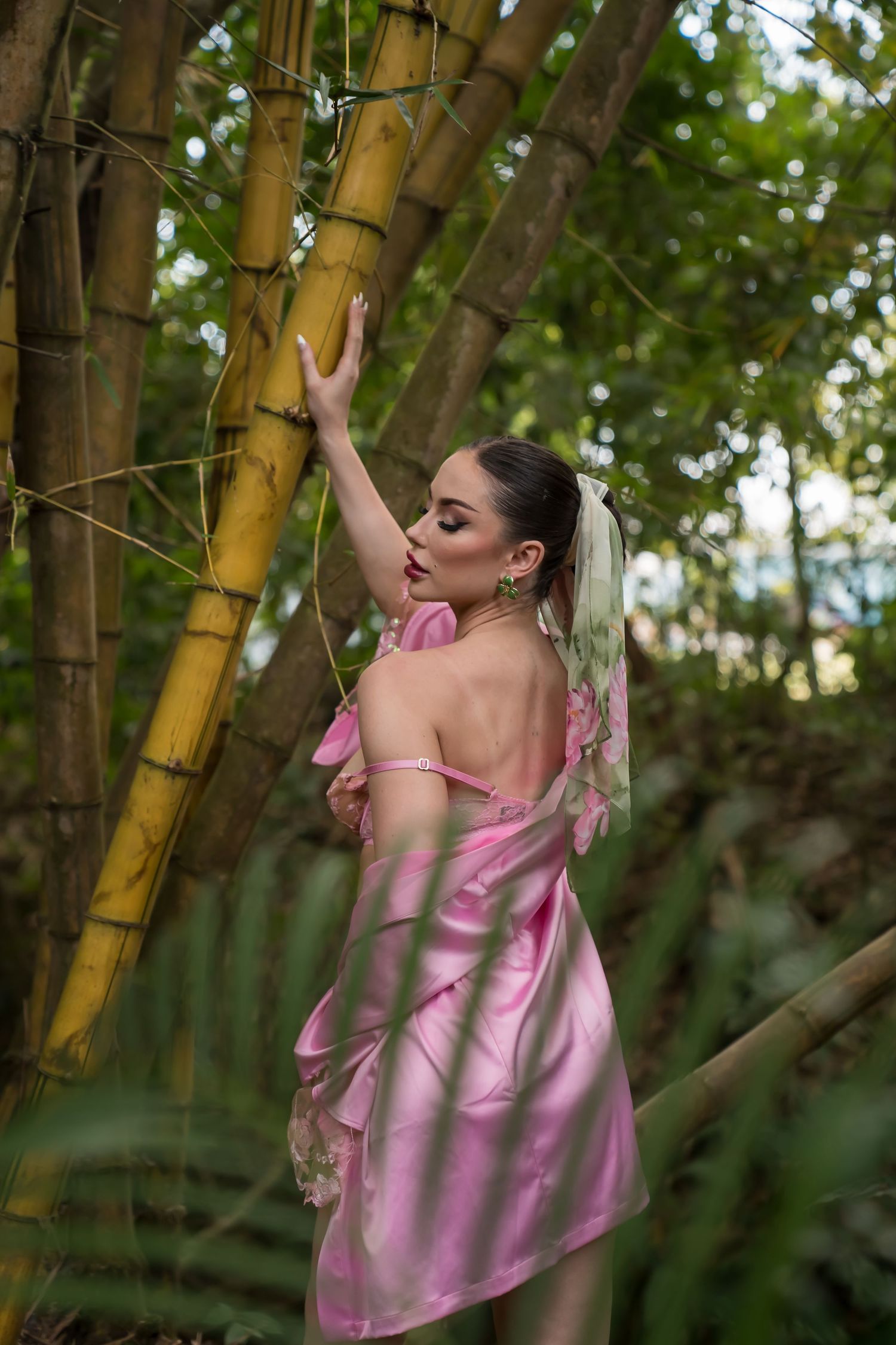 Woman in pink satin dress reaching up while posing elegantly among bamboo trees in a green forest.