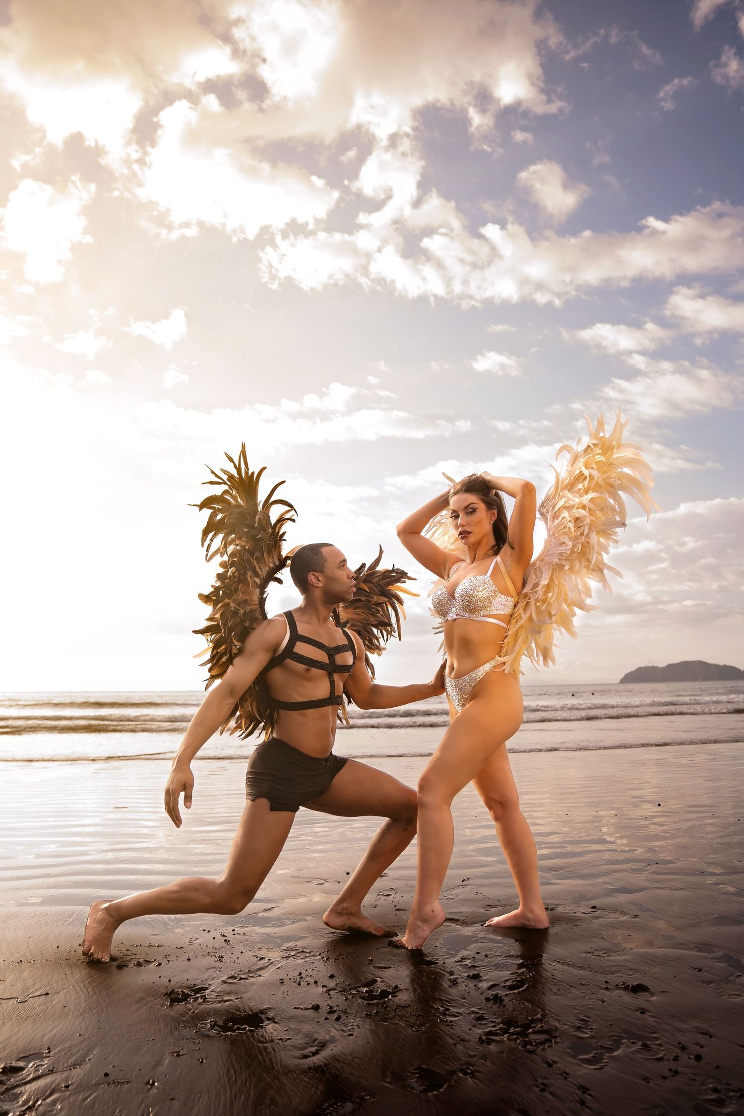 Two dancers in golden feathered costumes pose dramatically on a beach at sunset.
