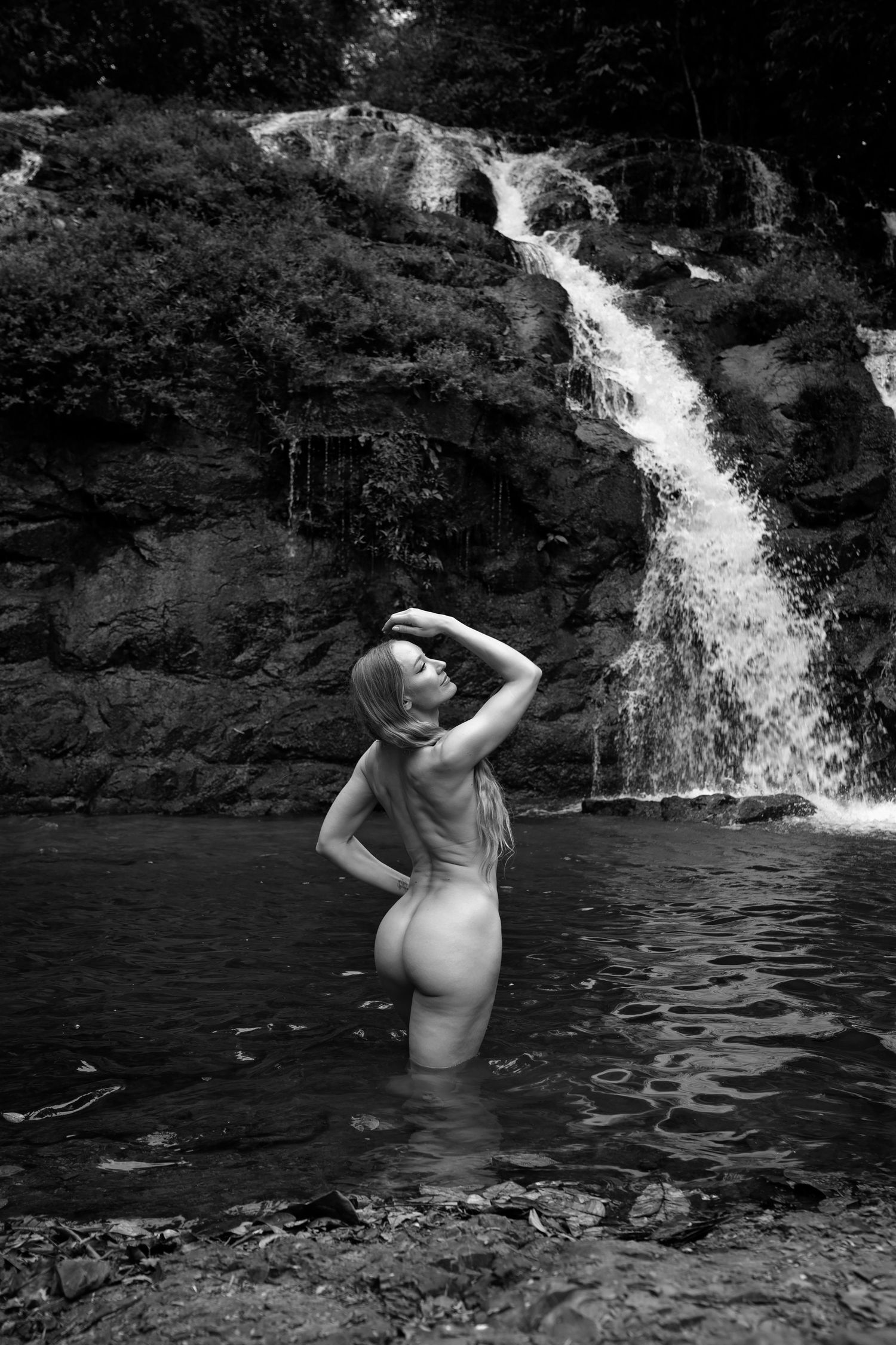 Black and white photo of a woman standing in a natural pool before a cascading waterfall surrounded by rocks.