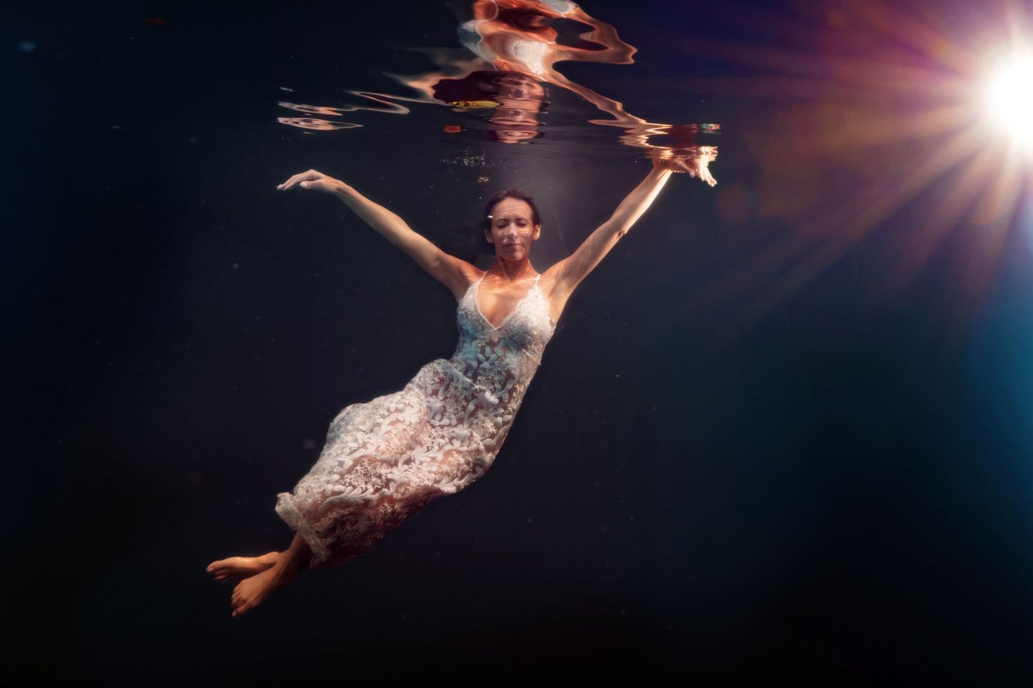 Woman in white dress floating gracefully underwater with sunlight streaming through the surface in a dark pool.