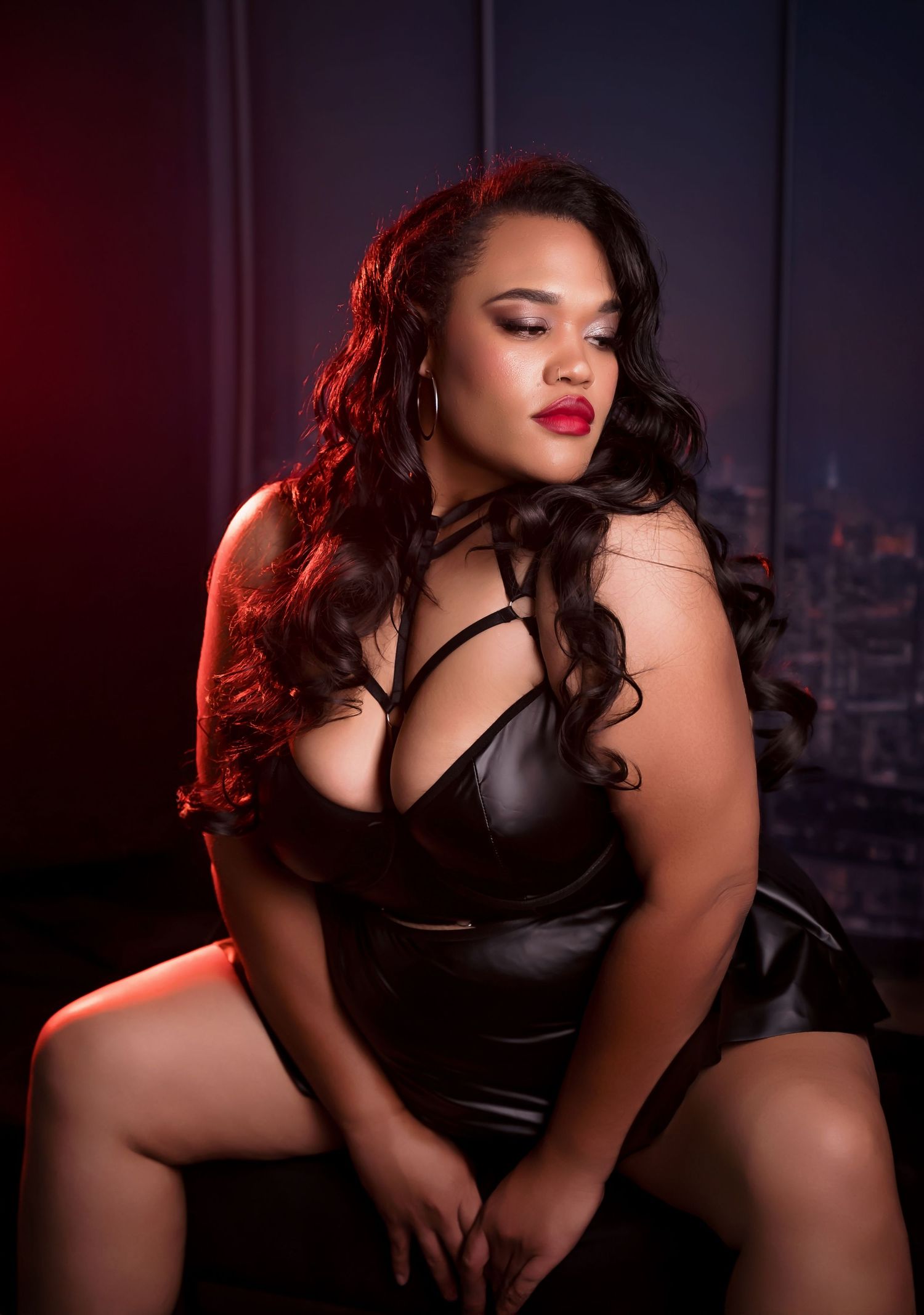 A confident plus-size woman poses in a black leather dress with strappy details, featuring red lips and long wavy hair.